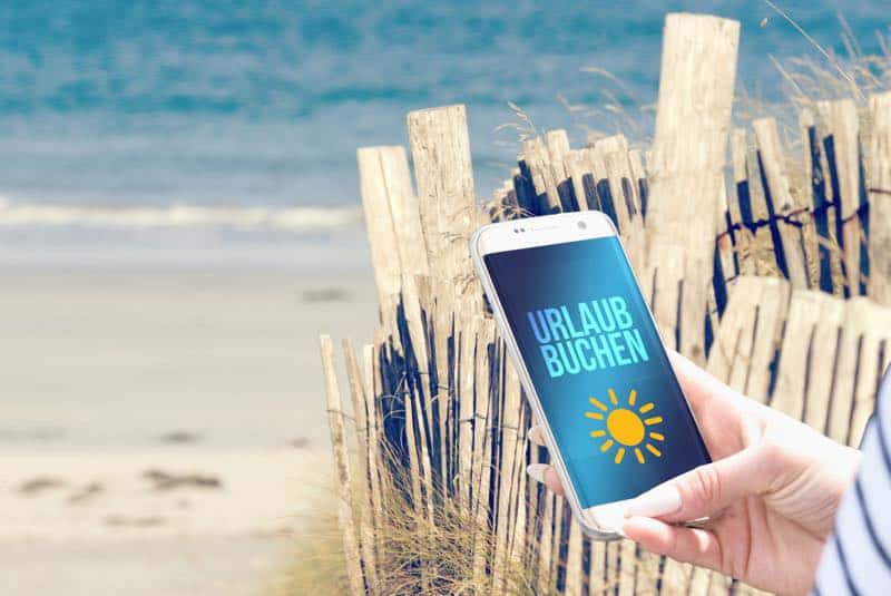 Smartphone am Strand mit Buchungs-App: Preise vergleichen und Urlaub clever buchen